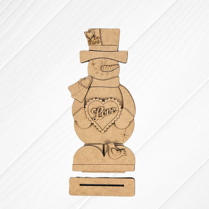 The Valentine Snowman Shelf Sitter by Janet's Craft Corner is a DIY decor craft kit that includes a wooden snowman with heart-shaped "Love" and "Be Mine" details, on a rectangular base with a small bird figure against a textured white and burlap background. Perfect for Valentine's Day decorating.