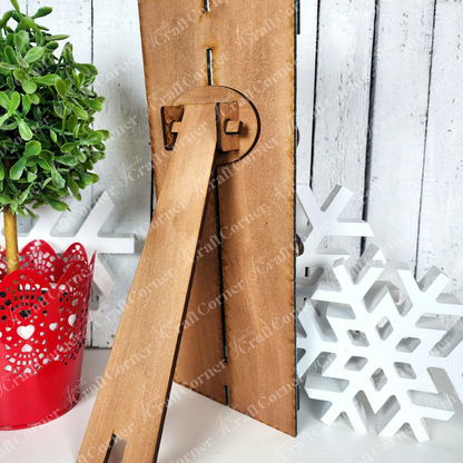 The Valentine Snowman Table Top Pallet Shelf Leaner by Janet's Craft Corner, a DIY seasonal home decor kit, is displayed on a white wooden surface beside a potted plant in a red holder and two white snowflakes—perfect for winter décor.