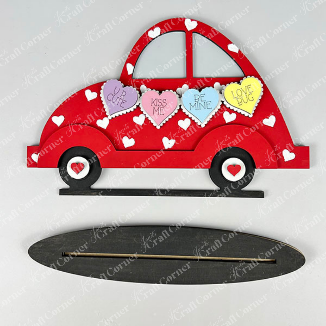 The Valentine Volkswagen Shelf Sitter by Janet's Craft Corner is a delightful red car-shaped decoration adorned with small white hearts and features four heart-shaped messages: "UR CUTE," "KISS ME," "BE MINE," and "LOVE BUG." It gracefully sits on a black oval base with a handy slot, ideal for displaying your handmade crafts.