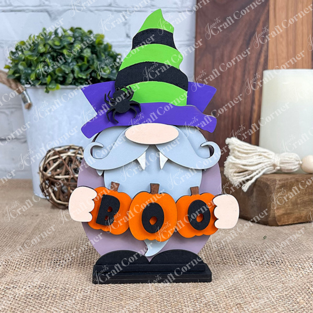A Vampire Gnome by Janet's Craft Corner, featuring a purple and green striped hat, a white beard, and a spider holding orange letters spelling "BOO," sits on burlap beside a potted plant and decorative tassels, adding the ideal touch of spooky charm against the stark white brick wall.