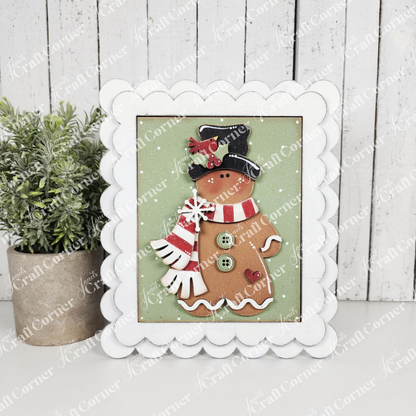 Framed artwork of a gingerbread man with a scarf on a wooden surface.