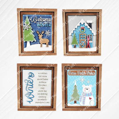 Displayed against a white wooden backdrop are four winter-themed decorations: Janet's Craft Corner's hand-painted Welcome Winter farmhouse signs featuring a deer, a polar bear, a framed list of winter words, and a house adorned with snow and skis. These items make for perfect home decor pieces.