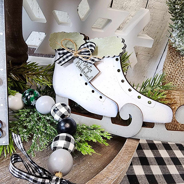 Displayed on a wooden surface, Janet's Craft Corner presents a decorative pair of white ice skates adorned with a green bow and a "Winter Wonderland" tag. This delightful Winter Wonderland Tiered Tray Set, encircled by green foliage and black-and-white checkered beads, makes for the perfect cozy home décor this winter.