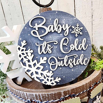 A charming home décor piece from the Winter Wonderland Tiered Tray Set by Janet's Craft Corner features a round rustic sign that rests on a wooden planter. It displays "Baby It's Cold Outside" in elegant white script over snowflakes and a snowy blue backdrop. A white snowflake ornament accompanies it, completing the winter-inspired ensemble.