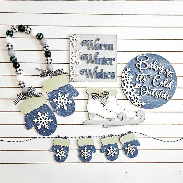 Winter-themed decorations displayed on a white wooden background include a "Warm Winter Wishes" sign, a "Baby it's Cold Outside" plaque, blue mittens adorned with snowflakes, beaded garland, and white ice skates. This captivating collection is reminiscent of the Winter Wonderland Tiered Tray Set by Janet's Craft Corner for cozy home décor.