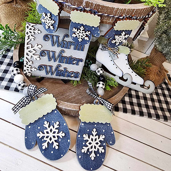 A cozy winter-themed display showcases the Winter Wonderland Tiered Tray Set from Janet's Craft Corner, featuring blue mittens, white snowflakes, and a "Warm Winter Wishes" sign. Complemented by decorative ice skates, greenery, and plaid fabric on a wooden surface, it makes the perfect DIY home décor for the season.