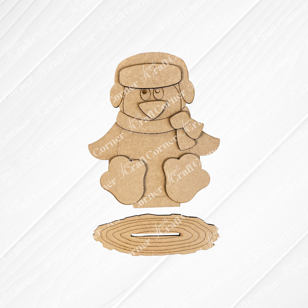 Janet's Craft Corner presents the Woodland Friends Penguin Shelf Sitter, a DIY home decor kit featuring a penguin in a scarf and winter hat, displayed against a white brick background. This charming kit includes arranged pieces for easy assembly.