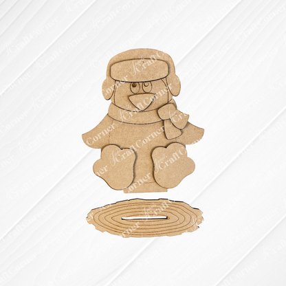 Janet's Craft Corner presents the Woodland Friends Penguin Shelf Sitter, a DIY home decor kit featuring a penguin in a scarf and winter hat, displayed against a white brick background. This charming kit includes arranged pieces for easy assembly.