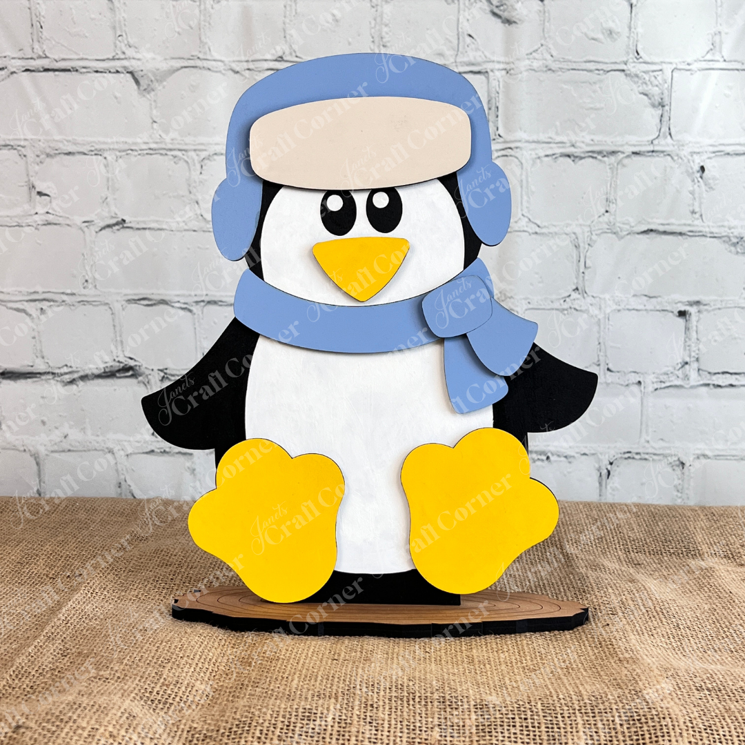 The Woodland Friends Penguin Shelf Sitter by Janet's Craft Corner features a cartoon penguin in a blue hat and scarf, yellow feet, leaning against a brick wall. 