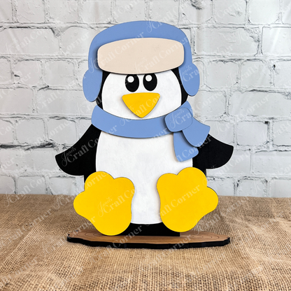 The Woodland Friends Penguin Shelf Sitter by Janet's Craft Corner features a cartoon penguin in a blue hat and scarf, yellow feet, leaning against a brick wall. 