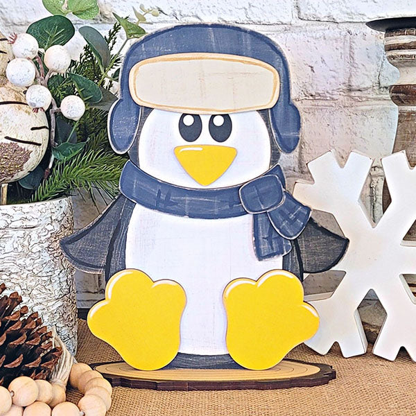 Add a touch of charm to your woodland friends-themed décor with Janet's Craft Corner's Woodland Friends Penguin Shelf Sitter. This adorable penguin, adorned with a scarf and hat, features yellow feet and beak as it sits on a wooden base. Surround it with pinecones, a large wooden snowflake, greenery, and white decorative berries for the perfect display.