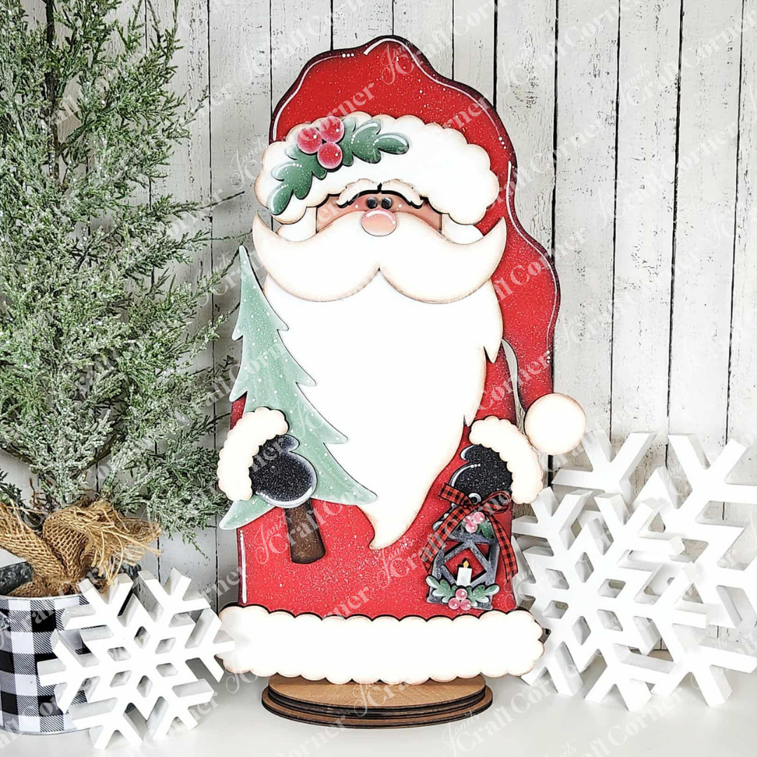 The Woodland Santa Shelf Sitter DIY craft kit by Janet's Craft Corner, holding a Christmas tree and lantern, sits between two white snowflake ornaments beside a green pine basket—festive décor on a white wood-paneled backdrop.