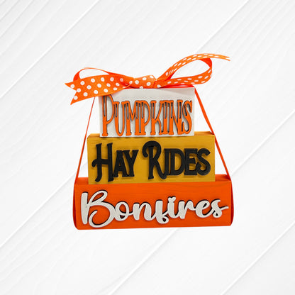 Add a festive touch with Janet's Craft Corner Word Stacks - Painted: stacked blocks reading "Pumpkins," "Hay Rides," and "Bonfires," finished with an orange polka-dot ribbon on a white wooden background.