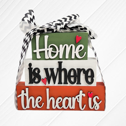 The Word Stacks - Painted by Janet's Craft Corner features three decorative wooden blocks in green, white, and red with "Home is where the heart is," red heart accents, and a black-and-white chevron ribbon on top.