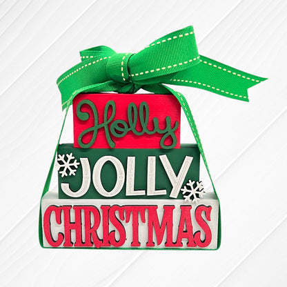 Janet's Craft Corner's Word Stacks - Painted is a festive ornament with "Holly Jolly Christmas" in red, green, and white, snowflake accents, a green ribbon bow on top, and set against a white wood background.
