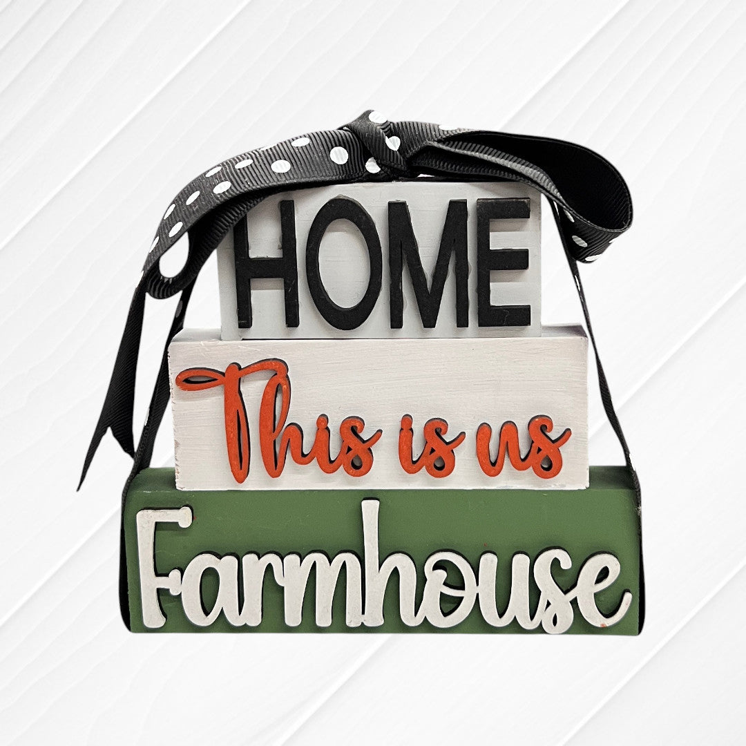 The Word Stacks - Painted by Janet's Craft Corner features three stacked wooden blocks reading "HOME," "This is us," and "Farmhouse" in black, orange, and white letters with a black polka dot ribbon on top. Background is white.
