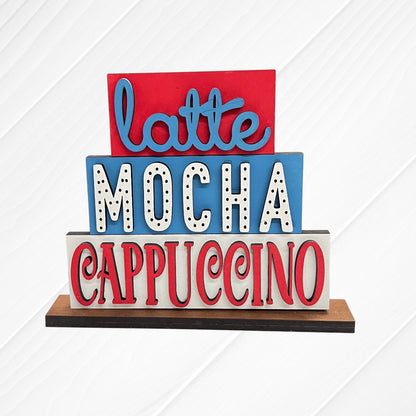 The Word Stacks - Painted by Janet's Craft Corner is a decorative sign with three stacked blocks featuring "latte," "mocha," and "cappuccino" in colorful, stylized fonts on a white, textured background.