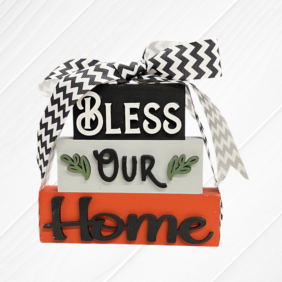 The Word Stacks - Painted by Janet's Craft Corner features three stacked wooden blocks labeled "Bless Our Home" in decorative letters, accented with green leaves and topped with a black and white chevron ribbon.