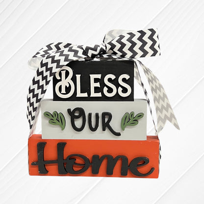 The Word Stacks - Painted by Janet's Craft Corner features three stacked wooden blocks labeled "Bless Our Home" in decorative letters, accented with green leaves and topped with a black and white chevron ribbon.