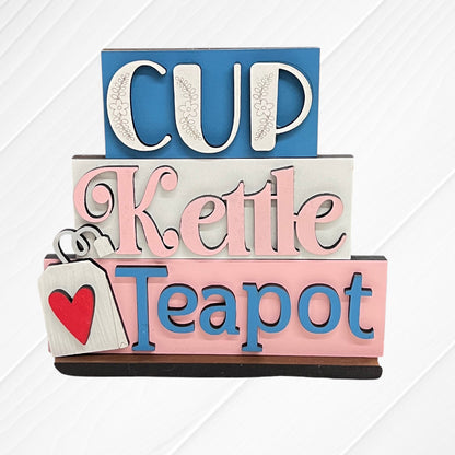 Janet's Craft Corner’s Word Stacks - Painted: a decorative sign with stacked words "Cup" (blue/white), "Kettle" (pink/white), and "Teapot" (blue/pink), plus a tea bag with a red heart, all on a white wood background.