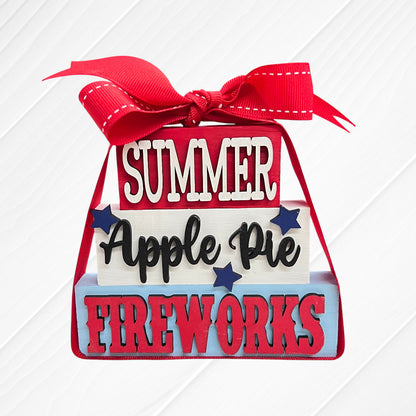 Janet's Craft Corner Word Stacks - Painted: Decorative blocks stacked with a red ribbon, featuring "SUMMER," "Apple Pie," and "FIREWORKS" text, blue star accents, on a white wood background.