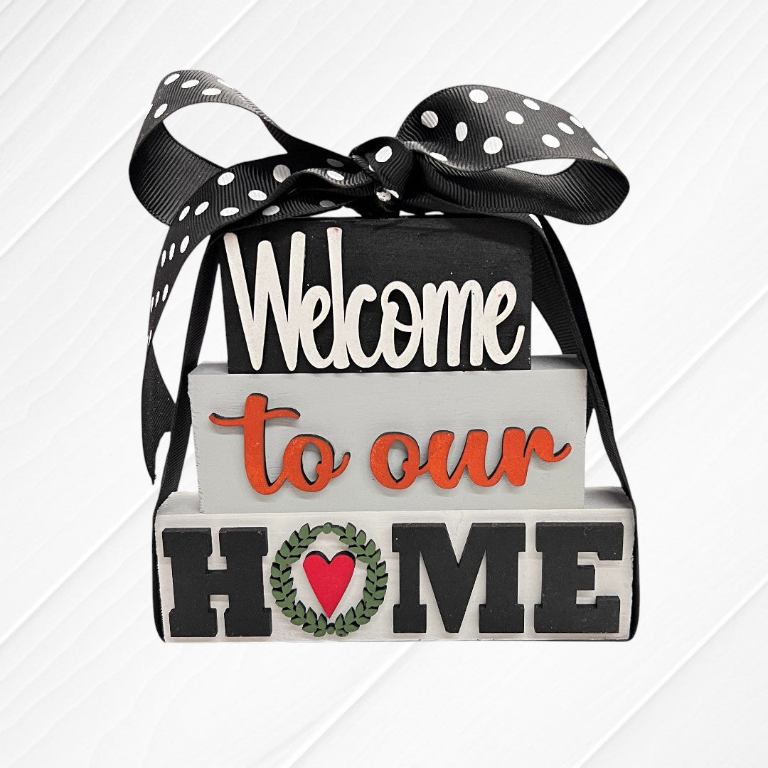 Janet's Craft Corner Word Stacks - Painted: Three stacked wooden blocks say "Welcome to our HOME," with a wreath and heart as the "O." Topped with a black and white polka dot ribbon, set against a white wood background.