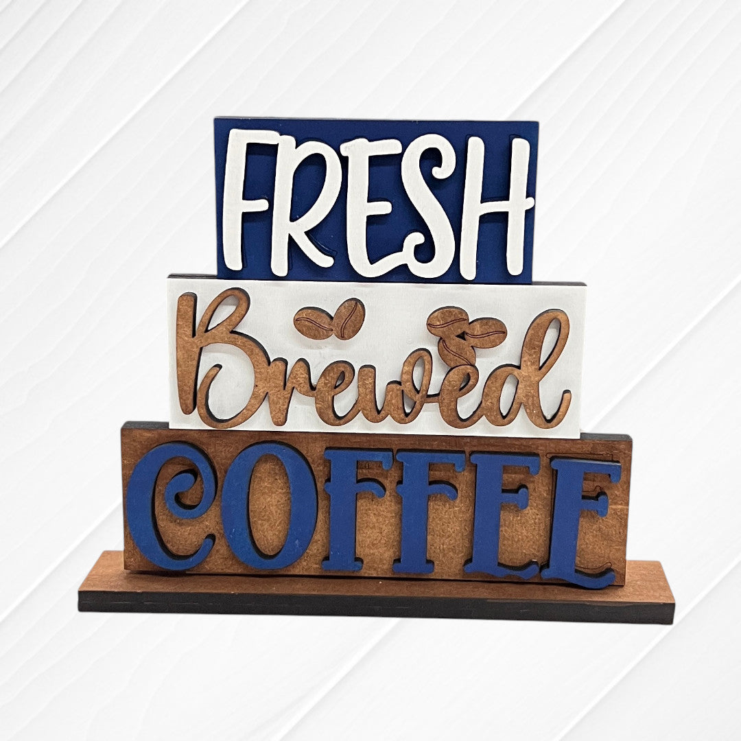 The Word Stacks - Painted sign by Janet's Craft Corner features three stacked wooden plaques reading “FRESH,” “Brewed” with coffee bean art, and “COFFEE” in bold blue/brown on a white textured background.