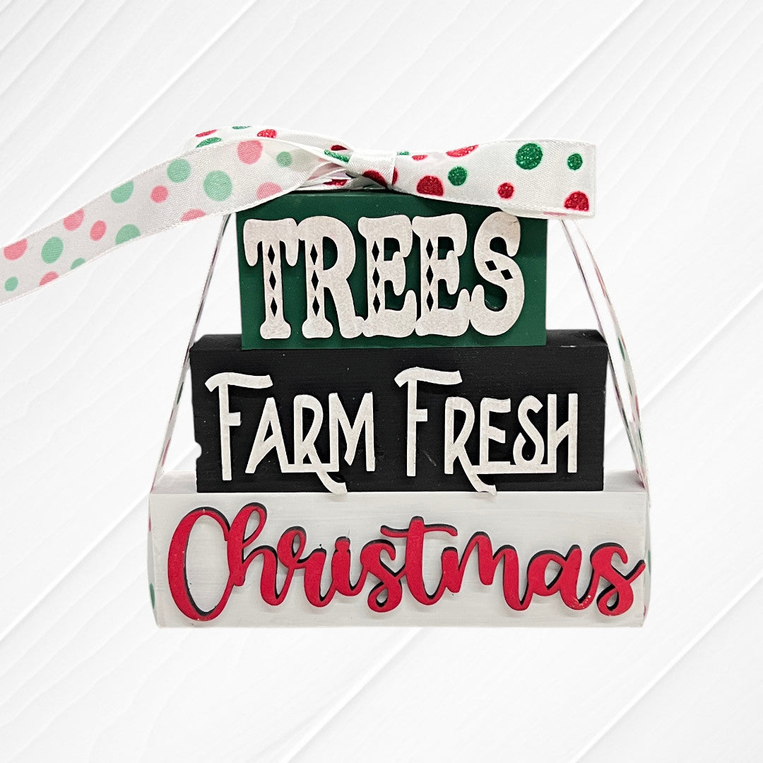 Janet's Craft Corner Word Stacks - Painted features three stacked wooden blocks reading "TREES," "Farm Fresh," and "Christmas," tied with a white ribbon with green, pink, and red dots on a white textured background.