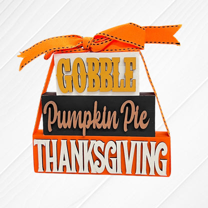 Janet's Craft Corner's Word Stacks - Painted is a decorative sign with an orange ribbon, featuring "GOBBLE" in yellow, "Pumpkin Pie" in tan script, and "THANKSGIVING" in bold white letters on orange.