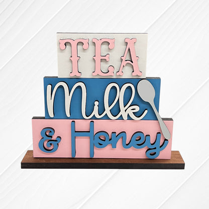 Janet's Craft Corner Word Stacks - Painted features stacked blocks reading "TEA," "Milk" with a spoon, and "& Honey" in pink and blue on a wooden base, set against a white textured background.