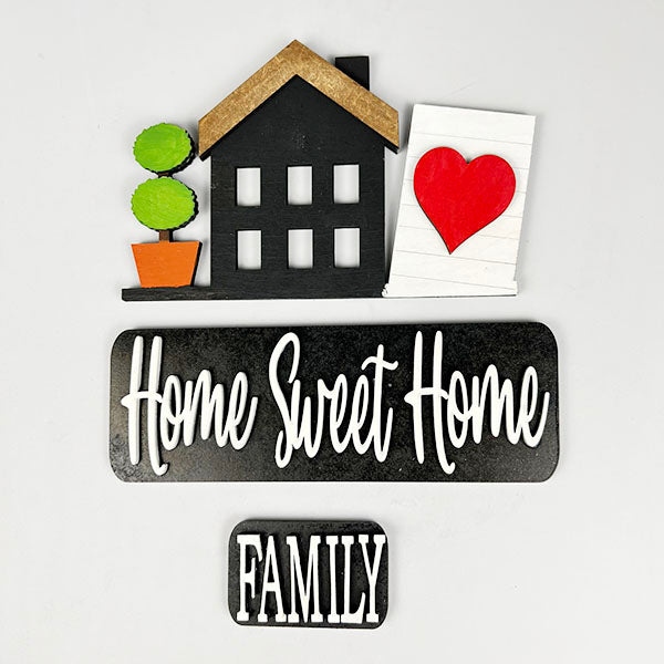 The Home Sweet Home Interchangeable Insert from Janet's Craft Corner showcases decorative wall art of a black house with a brown roof, accompanied by a potted plant and a white block adorned with a red heart for personalized charm. The elegant script reads "Home Sweet Home," while "Family" is prominently displayed in bold on a small black plaque, making it an ideal choice for versatile home decor.