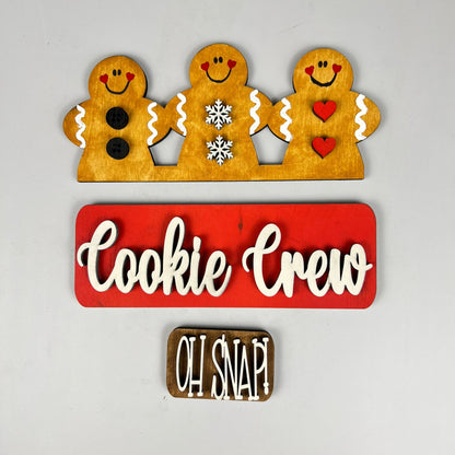 Three cheerful gingerbread figures—one with buttons, another with snowflakes, and the last with hearts—embellish a red sign that says "Cookie Crew," accompanied by a brown sign that reads "Oh Snap!" on a gray background. This charming DIY home decor kit from Janet's Craft Corner provides interchangeable decorations to brighten up the festive season.