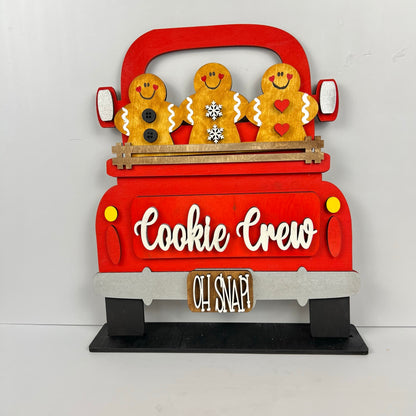 A delightful festive decoration from Janet's Craft Corner highlights three cheerful gingerbread figures riding in an antique truck labeled "Cookie Crew." Below, a small wooden sign reads "Oh Snap!" The truck, featuring white and brown accents, serves as the charming base for customizable inserts, offering a personalized touch. This is part of the Antique Truck Base with Interchangeable Insert DIY home decor kit.