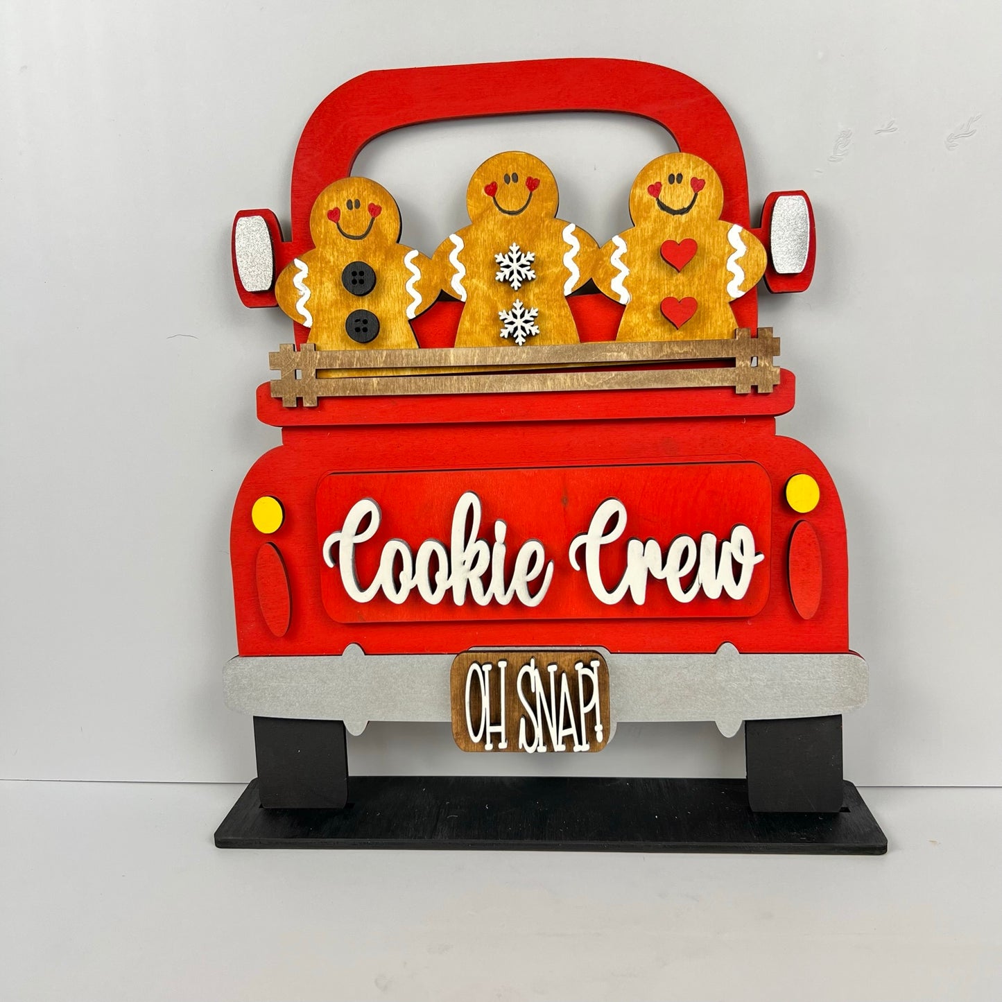 The Cookie Crew Interchangeable Insert from Janet's Craft Corner showcases three delightful gingerbread figures riding in a red truck labeled "Cookie Crew," with the whimsical phrase "Oh Snap!" underneath. Perfect as versatile decor, these gingerbread men are beautifully detailed with buttons, a snowflake, and hearts.