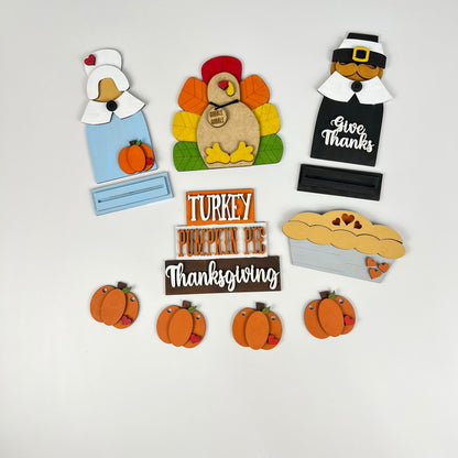 Introducing the Give Thanks/Thanksgiving Tiered Tray Kit by Janet's Craft Corner, a delightful addition to your Thanksgiving home décor. This festive arrangement features charming wooden decorations, including a pilgrim woman, a colorful turkey, and a pilgrim man with the phrase "Give Thanks." Complemented by a pie, pumpkins, and an additional sign reading "Turkey Pumpkin Pie Thanksgiving," all set against a crisp white background. Perfect for adding seasonal cheer to any tiered tray or table setting.