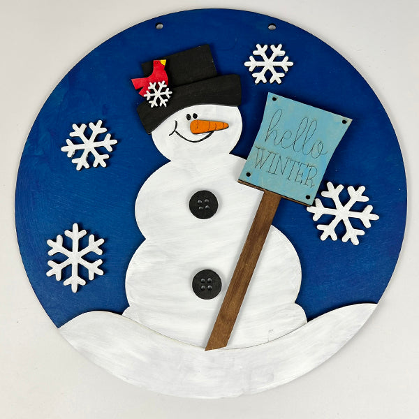 Embrace the beauty of winter with the "Hello Winter Door Hanger" from Janet's Craft Corner. This delightful DIY kit showcases a cheerful round snowman wearing a black hat with a red ribbon. He holds a light blue sign, set against a dark blue background decorated with white snowflakes—the perfect addition for seasonal charm.