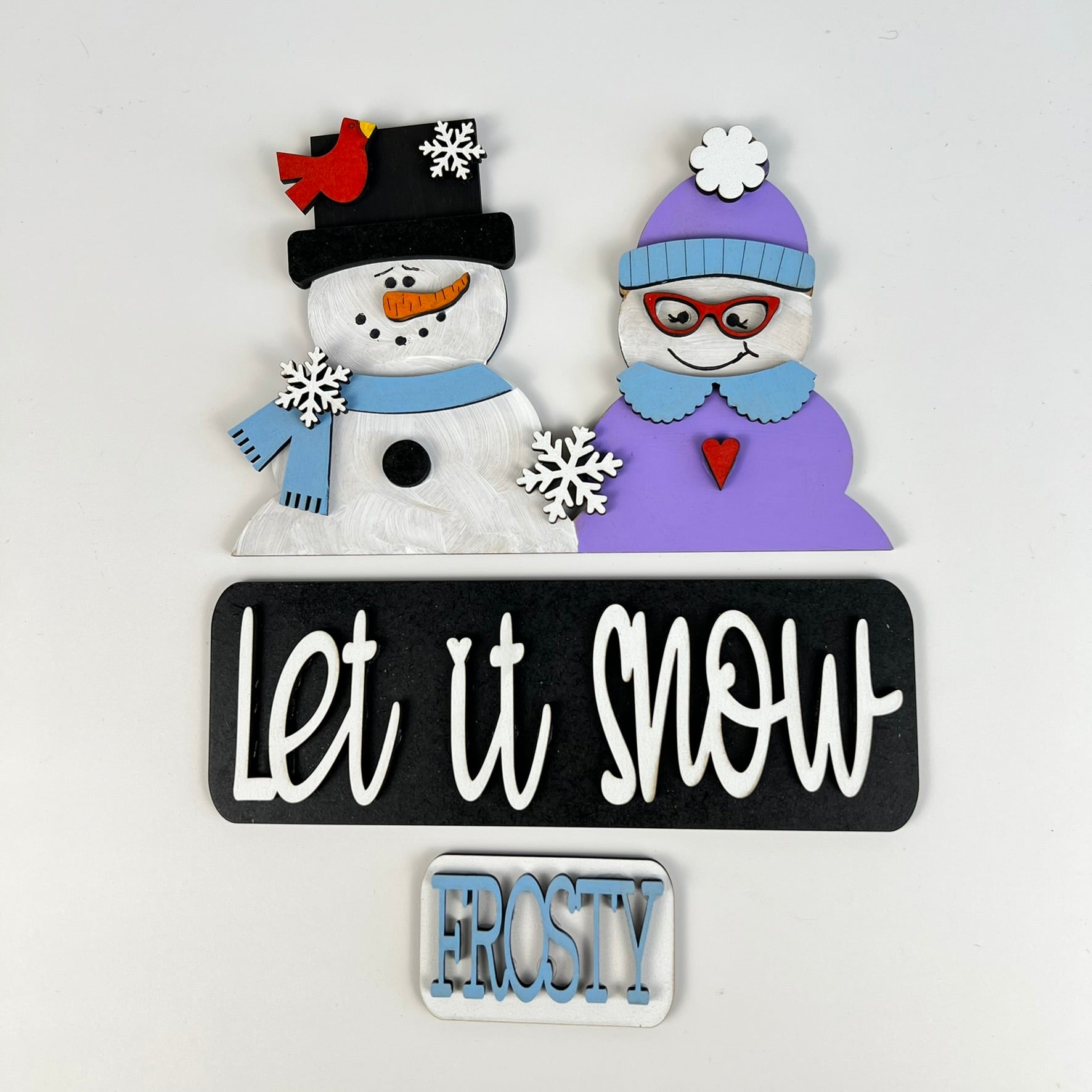 A charming decorative sign features a snowman couple: one wears a black hat with a carrot nose, while the other sports a purple hat and glasses. Snowflakes and a red bird float above them. The phrases "Let it Snow" and "Frosty" accompany the scene, making it an ideal addition for interchangeable craft inserts or your DIY home decor kit from Janet's Craft Corner, specifically the Let It Snow Interchangeable Insert.