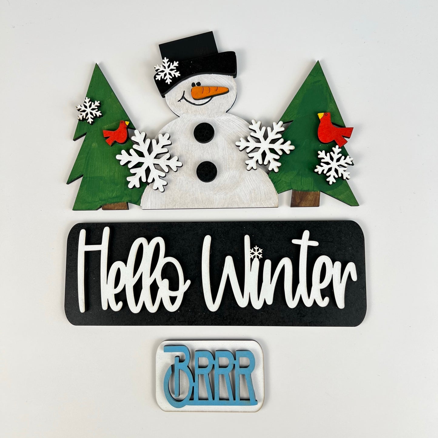 Transform your home this festive season with Janet's Craft Corner's "Hello Winter Interchangeable Insert - Painted." This winter-themed decor showcases a cheerful snowman adorned with a black hat and red scarf, set amidst vibrant green pine trees, charming red cardinals, and delicate white snowflakes. The design features a prominent black sign reading "Hello Winter" alongside a smaller "BRRR" sign, making it an ideal addition for interchangeable inserts on antique truck bases.