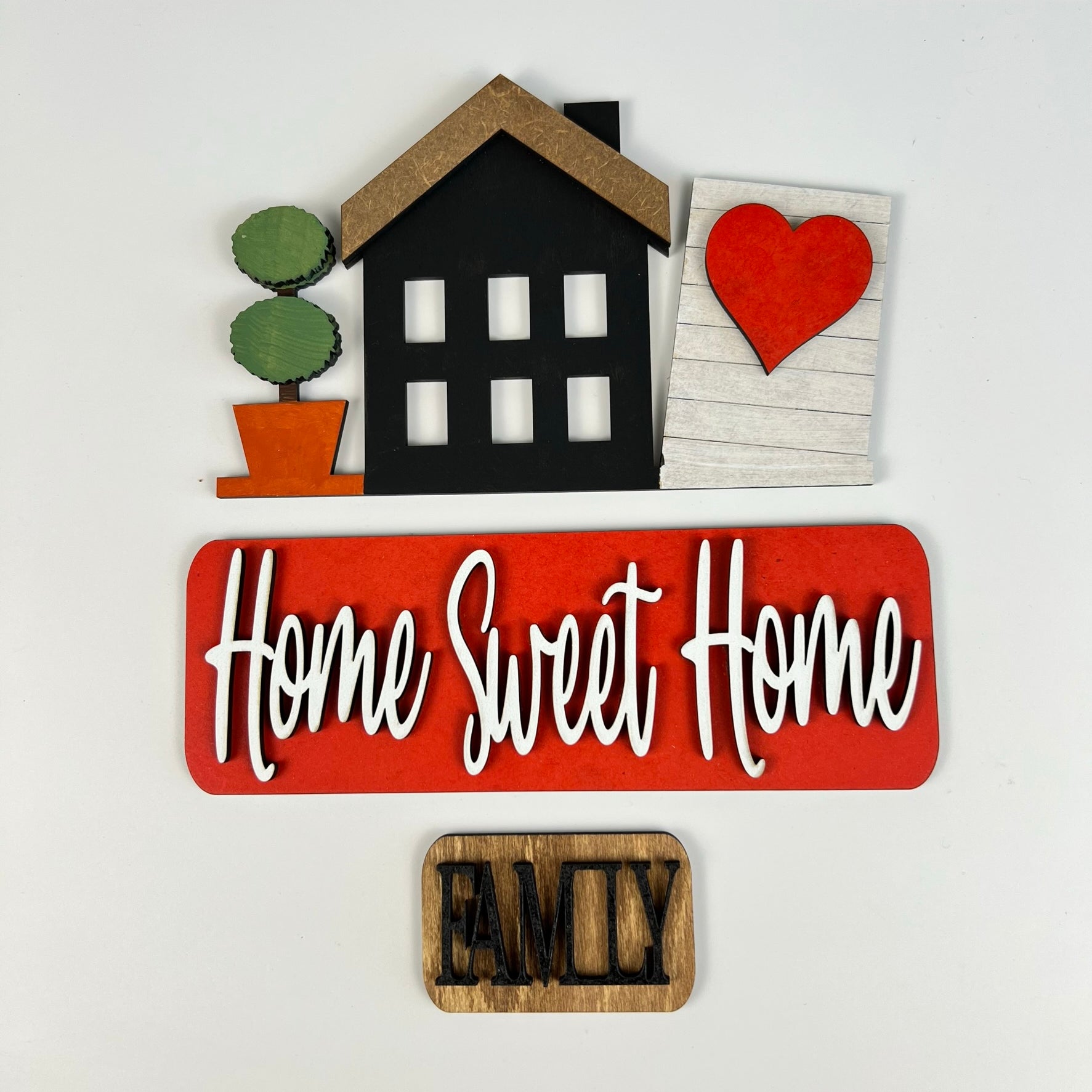 The Home Sweet Home Interchangeable Insert DIY home decor kit by Janet's Craft Corner features a black house, a potted plant, and a wooden board adorned with a red heart. The phrase "Home Sweet Home" is elegantly displayed in white on a red backdrop, complemented by the word "Family" on an interchangeable wood piece for personalized flair.