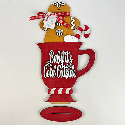 The "Gingerbread Hot Cocoa Mug Shelf Sitter" from Janet's Craft Corner features a cheerful red mug cutout with the phrase "Baby It's Cold Outside." This delightful decoration is adorned with a gingerbread hot cocoa figure holding a "Hot Cocoa 5¢" sign and a candy cane, completed by charming snowflake accents for that ideal festive atmosphere.