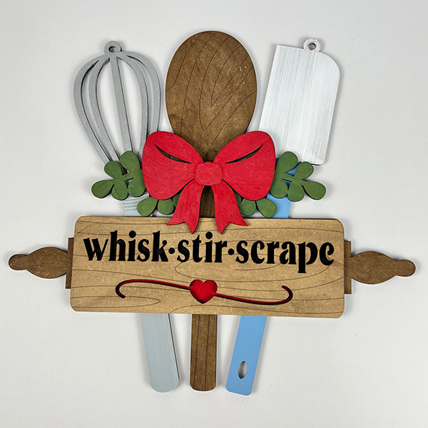 Introducing the "Whisk Stir Scrape Utensils Sign" by Janet's Craft Corner: This delightful decor set features kitchen utensil shapes adorned with a red bow, including a whisk, spoon, spatula, and rolling pin. Ideal for a DIY Door Hanger Kit or as charming Kitchen Utensils Decor. The rolling pin displays the words "whisk: stir: scrape" alongside a small red heart.