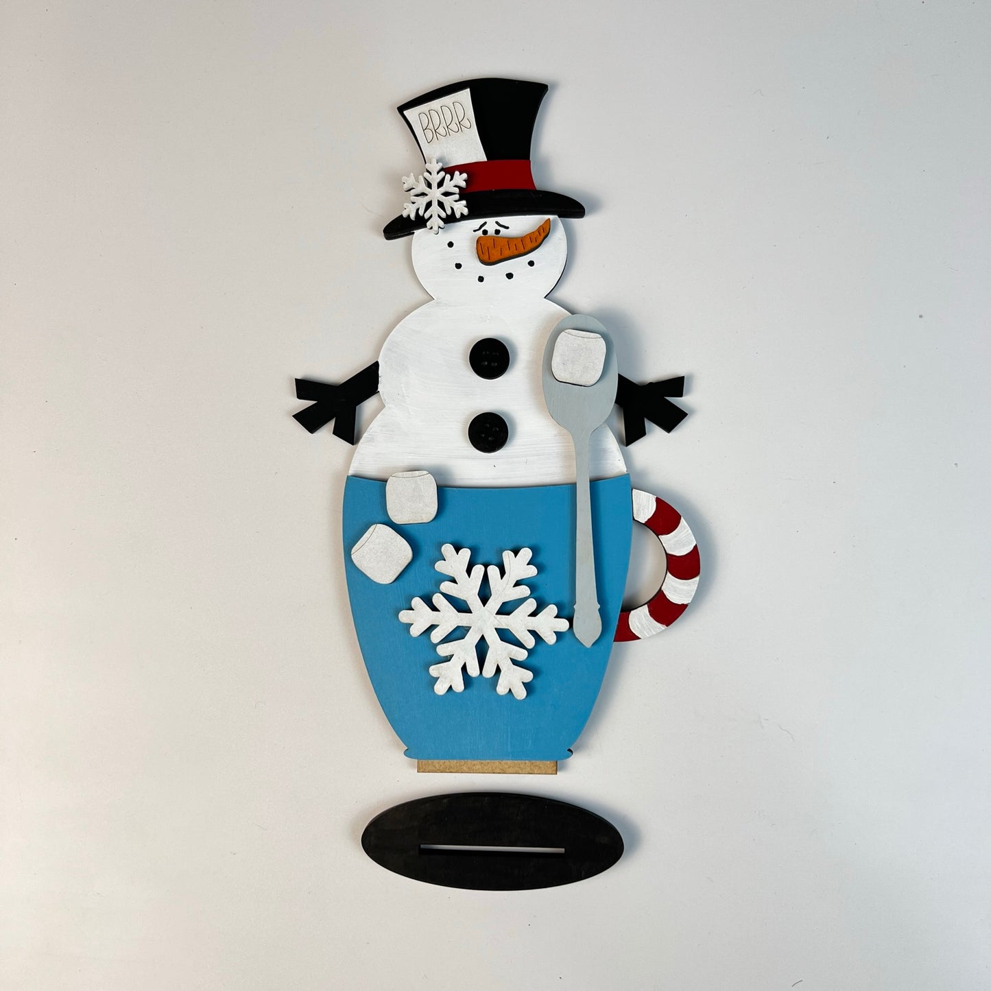 Introducing the Snowman Hot Cocoa Mug Shelf Sitter by Janet's Craft Corner. This delightful piece features a decorative wooden snowman dressed in a black top hat and scarf. The accompanying blue mug, adorned with white snowflakes, includes a charming candy cane handle. Ideal for home décor, it also comes with two marshmallows and a spoon for extra enjoyment.