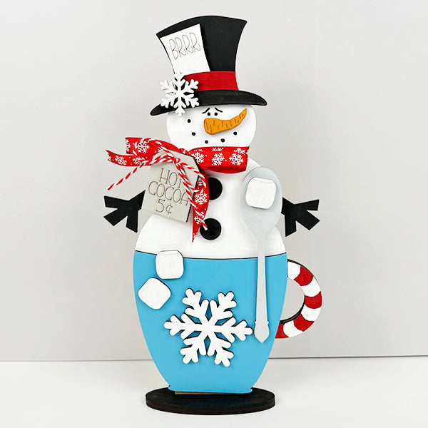 Decorate your home with the Snowman Hot Cocoa Mug Shelf Sitter by Janet's Craft Corner, showcasing a festive snowman sporting a black top hat, red scarf, and carrot nose in a blue mug adorned with a white snowflake. This delightful DIY kit is accentuated with marshmallows and a candy cane for the perfect holiday touch.