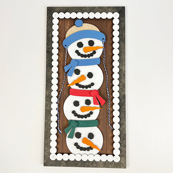 The Stacked Snowmen Shelf Leaner by Janet's Craft Corner features a vertical design with four smiling snowmen stacked atop one another, each adorned in scarves and hats of blue, red, and green. This charming holiday décor piece is enhanced with a border pattern of white circles, making it a delightful addition to any home décor collection.