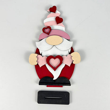 The Valentine Gnome Shelf Sitter by Janet's Craft Corner showcases a wooden design with a prominent white beard, pink and white striped hat, and red attire. Ideal for home décor, its hat and hands are adorned with red hearts while it holds a larger heart above the black rectangular base.