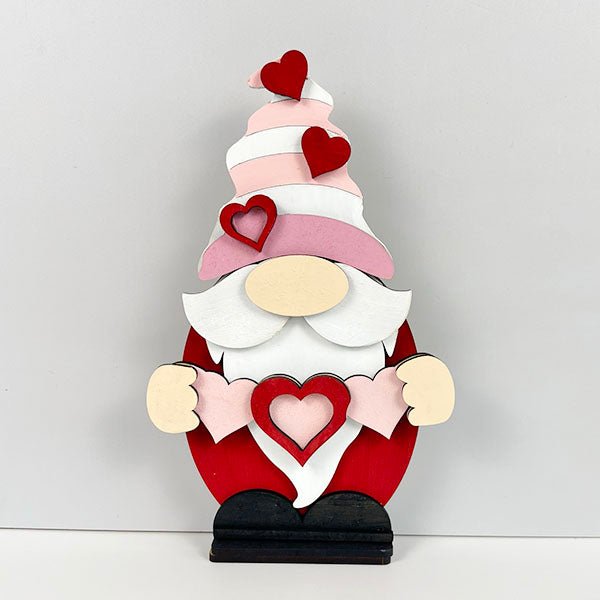 This delightful wooden figurine from Janet's Craft Corner, the Valentine Gnome Shelf Sitter, showcases a red body, white beard, and pink hat decorated with red hearts. It holds pink and red heart shapes and serves as an ideal piece of home décor against a light gray backdrop.