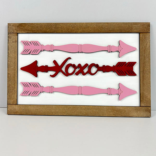 The Spindle Arrows Shelf Leaner from Janet's Craft Corner is a delightful home décor item featuring a wooden frame adorned with three horizontal arrows and the word "Xoxo" at the center. Ideal for DIY Kit enthusiasts, the design showcases pink arrows on the top and bottom, while the middle arrow matches the red "Xoxo," all set against a pristine white background.