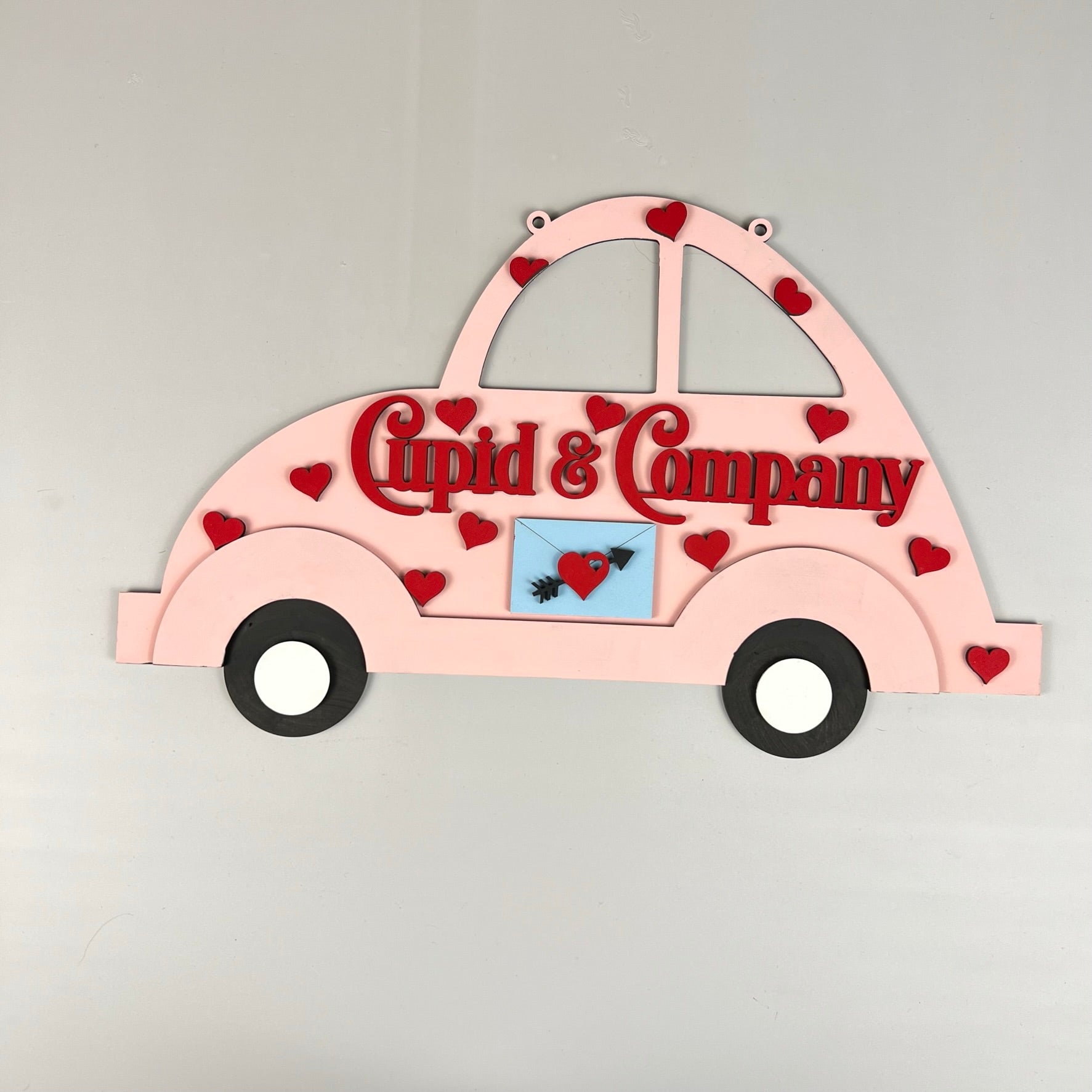 Introducing the Cupid Volkswagen Door/Wall Hanger from Janet's Craft Corner: This delightful home décor kit features a pink, car-shaped design known as "Cupid & Company." It is decorated with red hearts and a small image of a heart pierced by an arrow. The charming car comes complete with black wheels and white hubcaps, perfect for adding a touch of whimsy to any space.