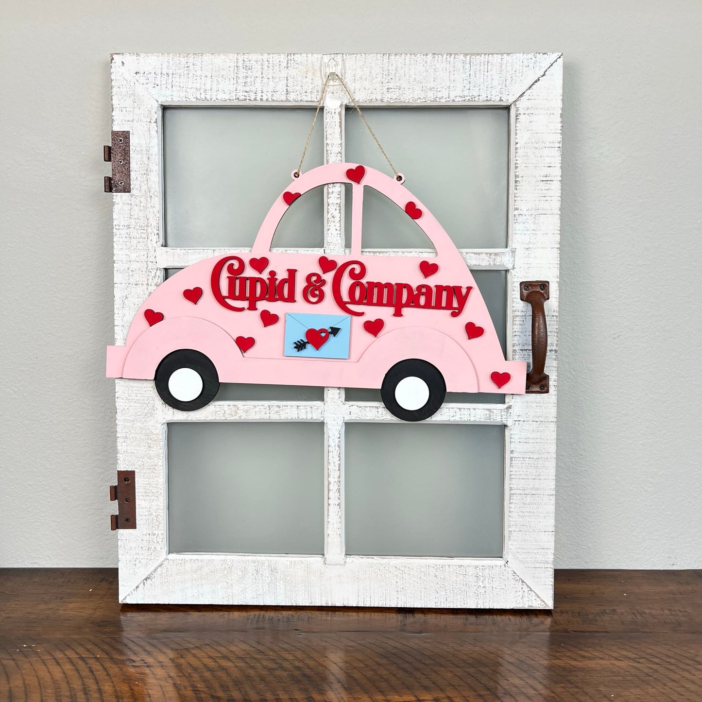 Introducing the Cupid Volkswagen Door/Wall Hanger by Janet's Craft Corner. This charming DIY decoration showcases a pink car cutout embellished with red hearts and the phrase "Cupid & Company." It elegantly hangs on a rustic white wooden window frame with frosted glass panels, making it an ideal choice for craft enthusiasts looking to enhance their home décor.