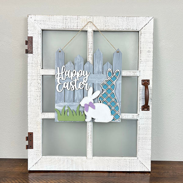 This delightful Easter Picket Fence Door Hanger from Janet's Craft Corner is a charming addition to your home décor. It features a decorative white wooden window frame with a rustic handle, showcasing bunnies and a fence. One bunny wears a purple bow, and the other displays a blue plaid pattern, with "Happy Easter" elegantly displayed on the sign.